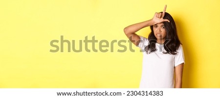 Image of sad african-american girl showing loser sign on forehead, feeling unconfident and lame, standing over yellow background.