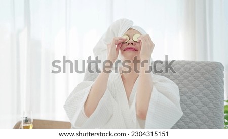 Young woman in bathrobe and towel turban putting fresh cucumber slices on eyes while relaxing on sofa bed at modern living room. Beauty spa concept Royalty-Free Stock Photo #2304315151
