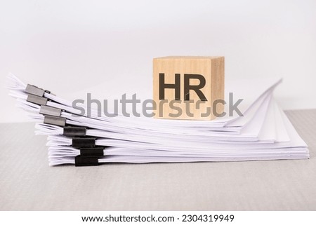wooden cube with text HR - acronym HUMAN RESOURCE MANAGEMENT - on a stack of documents. grey table, white background