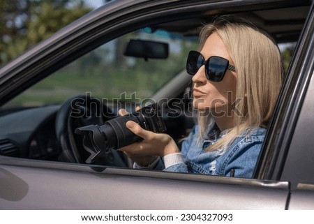 Paparazzi young woman in sunglasses sits in her car and takes pictures of famous person. Spy with camera in car.Private detective or paparazzi journalist sitting inside car,taking pictures with camera