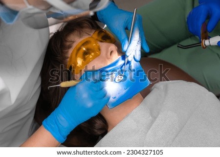 Applying resin based composite filling on tooth. Young woman at dental clinic. Female dentist with assistant treating cavities in patient mouth in modern dental office. Royalty-Free Stock Photo #2304327105