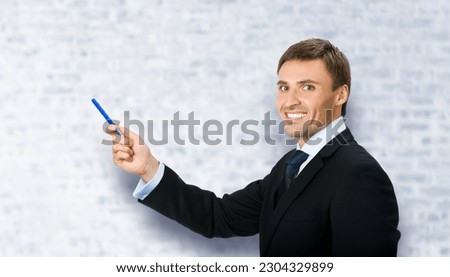 Image of business man in black confident suit, necktie, show pointing advertising product, on white bricks office background. Copy space, slogan text area.