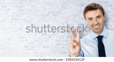 Excited businessman in black confident suit showing two fingers or victory sign gesture, on white bricks office background. Handsome happy man. Wide copy space for ad, slogan text.