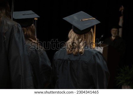 Unidentifiable graduates in black cap, gown, and golden tassel listen to a speech at a community college graduation ceremony. Royalty-Free Stock Photo #2304341465