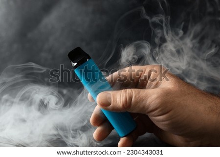disposable electronic cigarette in a man's hand on a dark background with smoke around. The concept of modern alternative smoking, vaping and nicotine Royalty-Free Stock Photo #2304343031