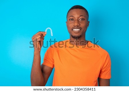 young man wearing orange T-shirt over blue studio background holding an invisible braces aligner, recommending this new treatment. Dental healthcare concept.