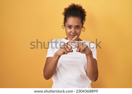 Young hispanic woman with curly hair standing over yellow background rejection expression crossing fingers doing negative sign 