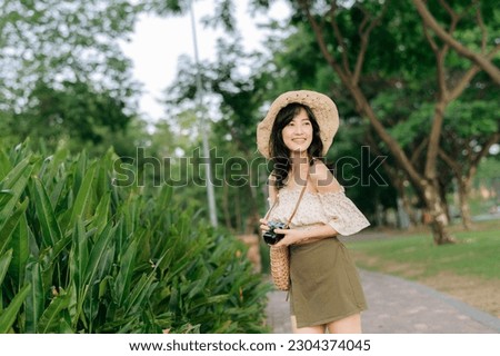 Portrait of young asian woman traveler with weaving hat and basket and a camera on green public park nature background. Journey trip lifestyle, world travel explorer or Asia summer tourism concept.