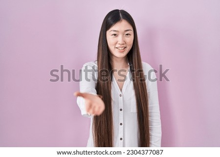 Chinese young woman standing over pink background smiling cheerful offering palm hand giving assistance and acceptance. 