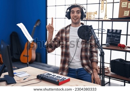 Young hispanic man playing piano at music studio pointing finger up with successful idea. exited and happy. number one. 