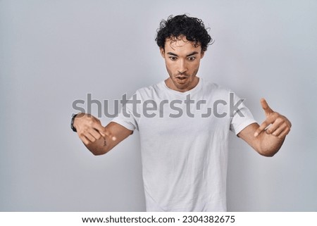 Hispanic man standing over isolated background pointing down with fingers showing advertisement, surprised face and open mouth 