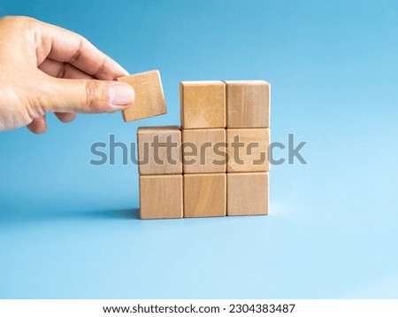 Last piece wood cube block in hand putting on wooden puzzles stacking arranging compleated isolated on blue background, minimal style. Solution, solve problem, business goal and success plan concepts. Royalty-Free Stock Photo #2304383487