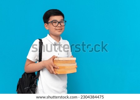 little asian schoolboy in glasses with backpack holding books and smiling on a blue background, korean child goes back to school and looks at copy space