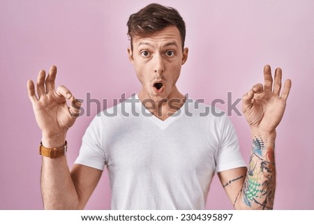 Caucasian man standing over pink background looking surprised and shocked doing ok approval symbol with fingers. crazy expression 