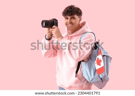 Young man with photo camera, backpack and flag of Canada on pink background