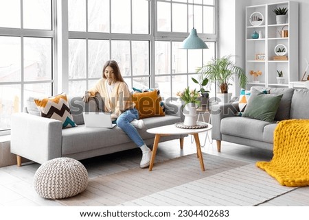 Beautiful young woman working with laptop on cozy sofa near big window Royalty-Free Stock Photo #2304402683