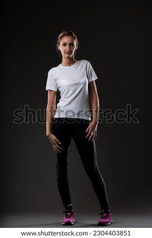 Blonde woman ready for workout, standing comfortably, hands on hips, looking at the camera. On black background, wearing dark tight pants, fuchsia sneakers, and a comfortable white t-shirt