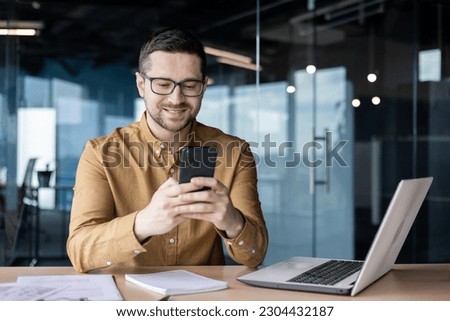 Smiling young male office worker sits at a desk with a laptop and writes a message on the phone, chats, checks mail, reads the news, break at work. Royalty-Free Stock Photo #2304432187