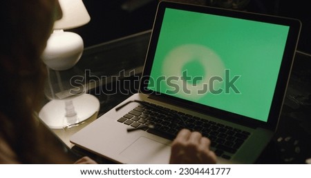 Over the shoulder shot of a business woman working in the evening in a room with the lights off on a computer on a table looking at a green screen. Using laptop computer with green laptop screen.