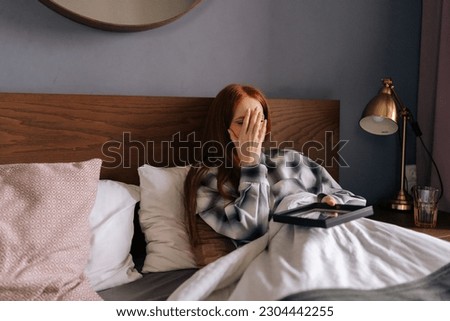 Depressed grief-stricken young woman lying on bed covering face with hand and crying while holding picture frame, touching photograph with love. Concept of nostalgia, grief, longing and loneliness.