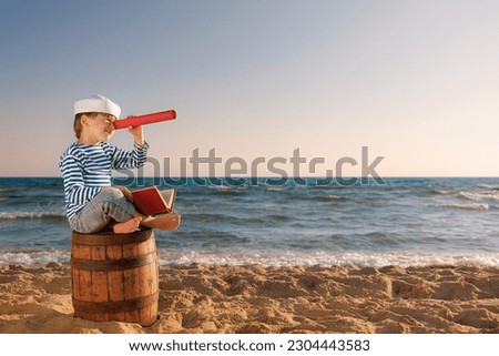 Child pretend to be sailor. Kid looking through spyglass. Child sitting on old barrel on the beach. Boy on summer vacation. Adventure and travel concept Royalty-Free Stock Photo #2304443583