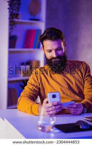 A cheerful bearded man is booking and paying for a trip on the phone while sitting at the neon-lighted home office and smiling at the phone. A hipster is using an online mobile app for booking.