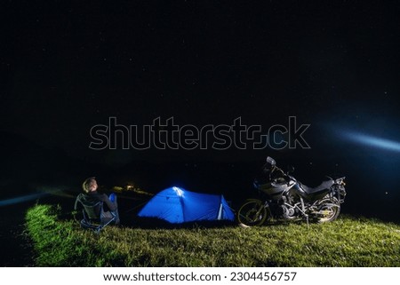 A man sits on a folding tourist chair and enjoys the view. Camping on top of a mountain. The stars are shining. A tent and a touring motorcycle. Travel and extreme recreation. Ukraine