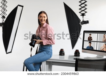 Happy creative photographer lady sitting on table workplace, holding her DSLR camera, smiling at camera while working with computer in modern photostudio