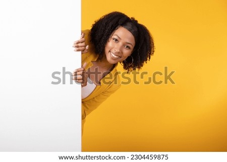 Intresting offer. Happy black lady peeping out the side of white advertisement board for your text or design. Excited woman standing near billboard over yellow background Royalty-Free Stock Photo #2304459875