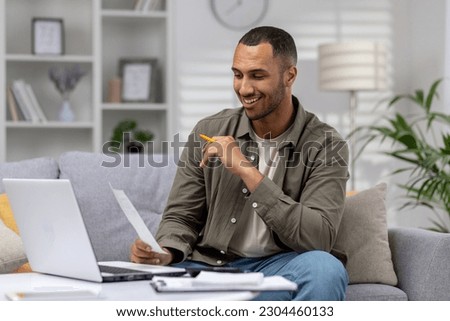 Young successful hispanic man at home sitting on sofa in living room, calculating and paying bills, man with calculator and laptop online paying for rent and loan. Royalty-Free Stock Photo #2304460133