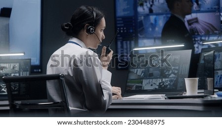 Female security officer works on computer in police monitoring center with CCTV cameras video footage. Male worker controls surveillance cameras with AI face scanning system on big digital screen. Royalty-Free Stock Photo #2304488975