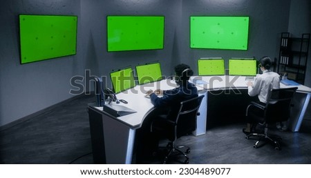 Multiracial male and female officers control CCTV cameras, work in security control center. Big digital screens and computer monitors with green screen, showing surveillance cameras video footage.