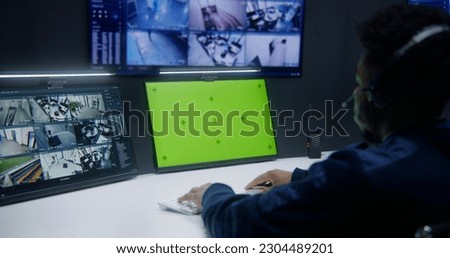African American male officer controls CCTV cameras in security monitoring center. Computer monitor with chroma key. Worker and big digital screen with surveillance cameras video footage on background