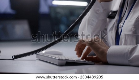 Female security operator works in police monitoring center. Woman types on keyboard, uses landline phone and walkie talkie. Computer and digital tablet screens with CCTV cameras footage on background.