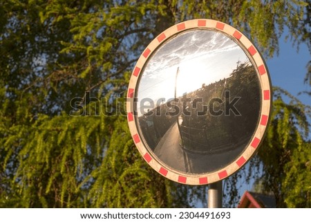 A mirror road sign at a crossroads in the suburbs. Road mirror against the background of trees. Reflected in the mirror : houses , street and setting sun . 