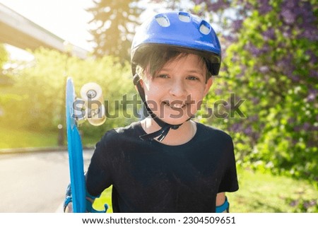portrait of a boy in a purple safety helmet, holds a blue plastic city cruiser, skateboard, in a sunny park in summer