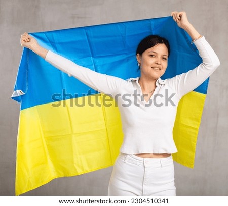 Positive middle aged woman in casual wear holding an unfurled flag of Ukraine in her hands raised above her head against a gray wall, studio shot. Support, national identification