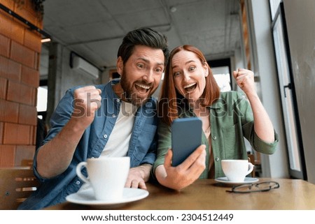 Overjoyed couple of friends holding mobile phone shopping online with sales, win money, celebration success sitting in modern cafe. Happy successful freelancers receive payment, reading good news  Royalty-Free Stock Photo #2304512449