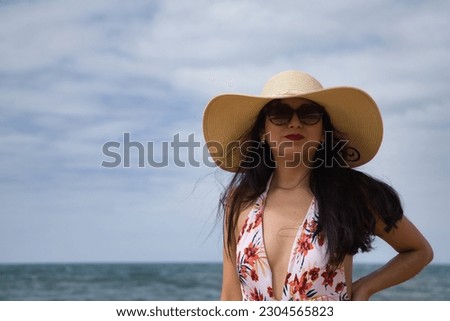 South American woman, young and beautiful, brunette with sunglasses, hat and swimsuit posing happy and smiling on the beach. Concept sea, sand, sun, beach, vacation, travel, summer.