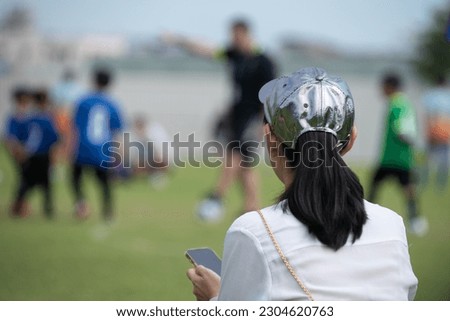 Mom sitting and watching her son playing football in a school football tournament on a sideline with a sunny day. Sport, outdoor active, lifestyle, happy family and soccer mom and soccer dad concept. Royalty-Free Stock Photo #2304620763