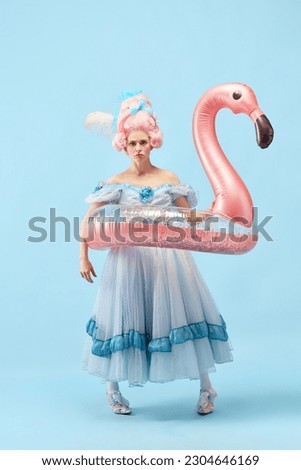 Portrait with funny, upset princess, queen wearing beautiful dress holding pink inflatable flamingo and going to swim over blue background. Concept of comparison of eras, vacation, summer, weekend Royalty-Free Stock Photo #2304646169