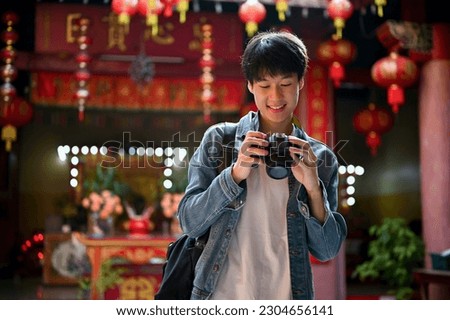 Happy and handsome young Asian male traveler is checking his pictures on his camera while visiting a beautiful Chinese's temple. Solo travel trip concept