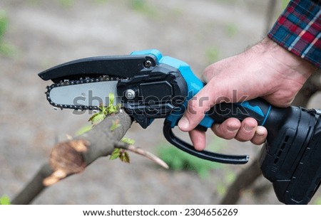 A hand holds a mini chainsaw with a battery to cut a broken tree branch on a sunny day. Royalty-Free Stock Photo #2304656269