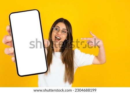 Mobile offer, excited caucasian lady pointing finger to smartphone in her hand for great mobile offer. Looking mobile phone with empty blank screen mockup. Isolated orange studio background.