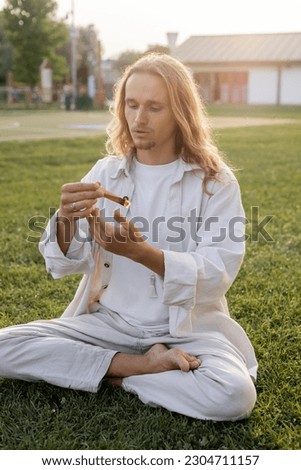 young long haired man in white linen clothes sitting in easy pose and holding scented palo santo stick on green lawn