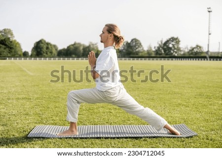 side view of barefoot man practicing yoga in warrior pose with anjali mudra gesture on green grass of outdoor stadium Royalty-Free Stock Photo #2304712045