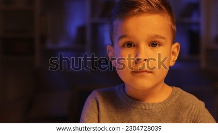 Portrait of a cute little boy sitting in his room in the evening looking at the camera. Close view. Royalty-Free Stock Photo #2304728039