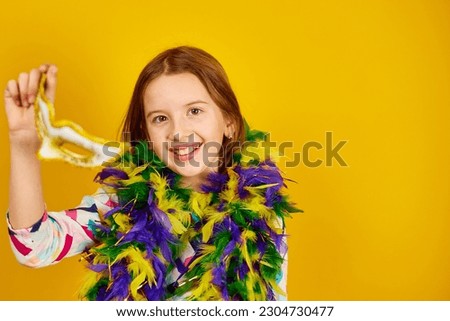 A joyful teenager girl wearing a colorful Brazil carnival mask, posing on yellow studio background, ready for celebration, birthday party, holidays concept