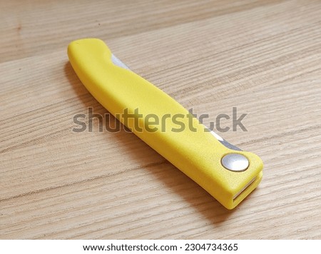 A folding kitchen knife with a yellow plastic handle and a serrated blade lies on a wooden board. Close-up.