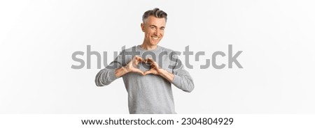 Portrait of cheeky middle-aged man in grey sweater, smiling and showing heart sign, love someone, standing over white background.